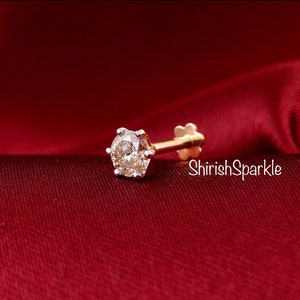 May include: A gold nose stud featuring a round, clear, faceted gemstone held in a six-prong setting. The post is straight with a flat, flower-shaped backing. The text ShirishSparkle is visible in white script lettering next to the jewelry piece.