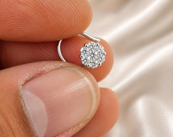 May include: A silver nose ring featuring a cluster of small, sparkling clear gemstones arranged in a floral pattern. The nose ring is held between two fingers against a soft, off-white background.