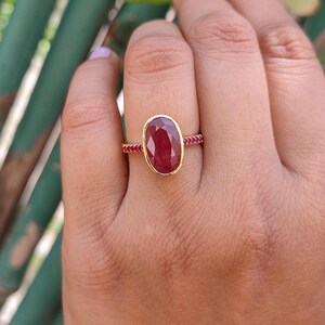 K&ouml;nnte beinhalten: Ein Goldring mit einem gro&szlig;en, oval geschliffenen, tiefroten Edelstein in einer glatten Fassung. Die Ringschiene ist schmal und mit einer Reihe kleiner, runder, leuchtend roter Steine verziert, die sich entlang der Seiten des Rings erstrecken.