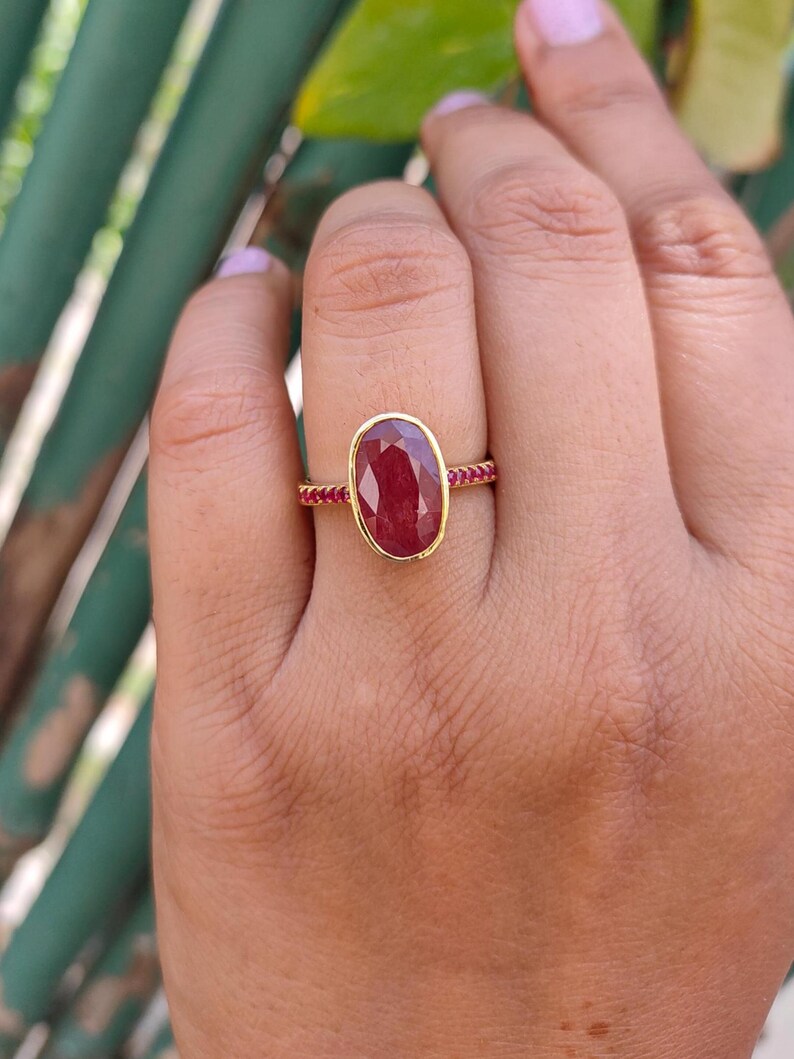 K&ouml;nnte beinhalten: Ein Goldring mit einem gro&szlig;en, oval geschliffenen, tiefroten Edelstein in einer glatten Fassung. Die Ringschiene ist schmal und mit einer Reihe kleiner, runder, leuchtend roter Steine verziert, die sich entlang der Seiten des Rings erstrecken.