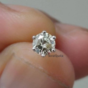 May include: A round brilliant cut gemstone stud earring set in a six-prong silver-toned metal setting. The stone features multiple facets that reflect light, and the text ShirishSparkle is printed in small black letters near the base of the setting.