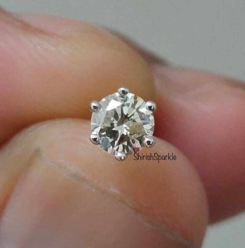 May include: A round brilliant cut gemstone stud earring set in a six-prong silver-toned metal setting. The stone features multiple facets that reflect light, and the text ShirishSparkle is printed in small black letters near the base of the setting.