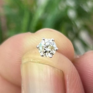 May include: A round brilliant cut gemstone set in a silver-colored metal six-prong stud setting. The stone features multiple facets that reflect light, creating a clear, sparkling appearance. The setting includes a thin metal post extending from the back.