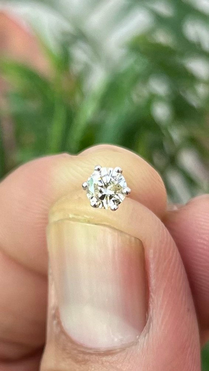 May include: A round brilliant cut gemstone set in a silver-colored metal six-prong stud setting. The stone features multiple facets that reflect light, creating a clear, sparkling appearance. The setting includes a thin metal post extending from the back.