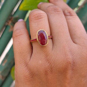K&ouml;nnte beinhalten: Ein Goldring mit einem gro&szlig;en, ovalen, tiefroten facettierten Edelstein in einer glatten Fassung. Die Ringschiene ist mit einer Reihe kleiner, runder, leuchtend roter Steine verziert, die beidseitig in das Band eingelassen sind.