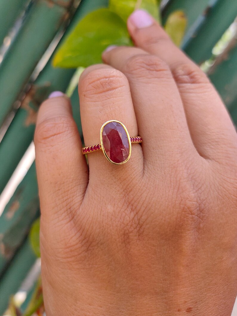 K&ouml;nnte beinhalten: Ein Goldring mit einem gro&szlig;en, ovalen, tiefroten facettierten Edelstein in einer glatten Fassung. Die Ringschiene ist mit einer Reihe kleiner, runder, leuchtend roter Steine verziert, die beidseitig in das Band eingelassen sind.