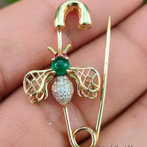 May include: A gold-coloured bee-shaped brooch with a green gemstone body and a diamond-studded abdomen. The brooch is attached to a safety pin design. The wings are detailed with a delicate openwork pattern. The text "Shirish Sparkle" is visible.