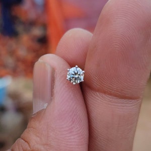 May include: A small, clear, round diamond gemstone set in a silver setting. The diamond is held in a person's hand.