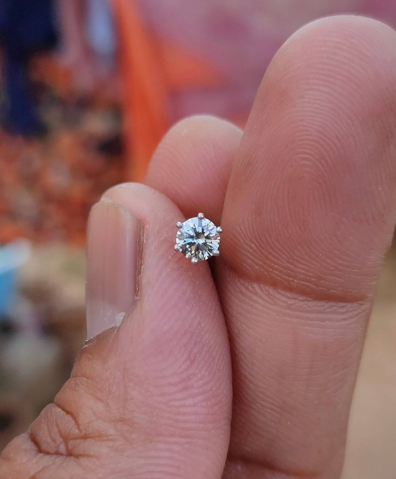 May include: A small, clear, round diamond gemstone set in a silver setting. The diamond is held in a person's hand.