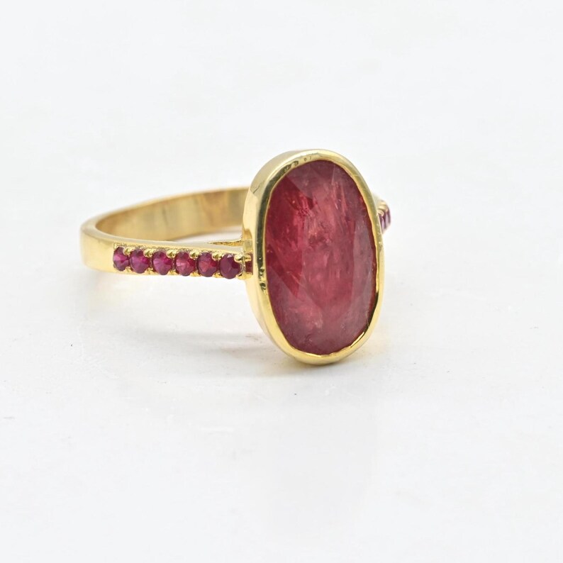 K&ouml;nnte beinhalten: Ein Goldring mit einem gro&szlig;en, ovalen, dunkelrosa Edelstein in einer Fassung. Das Band ist mit einer Reihe kleiner, runder, leuchtend rosa Steine verziert, die in die Seiten eingelassen sind und ein detailliertes Schmuckst&uuml;ck bilden.