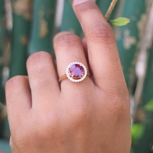 May include: A ring with a large, round, red gemstone surrounded by a halo of smaller, white gemstones. The ring is set in gold.