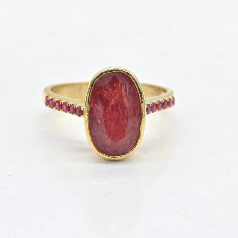 K&ouml;nnte beinhalten: Ein Goldring mit einem gro&szlig;en, ovalen, facettierten roten Edelstein in einer Zargenfassung. Die Ringschiene ist auf beiden Seiten mit einer Reihe kleiner, runder, roter Steine besetzt, die von der zentralen Fassung ausgehen.