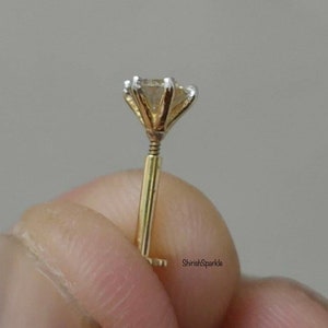 May include: A gold nose stud featuring a single round clear gemstone held in a six-prong setting. The post is straight with a threaded screw mechanism near the base of the setting. The text ShirishSparkle is visible on the lower right side.