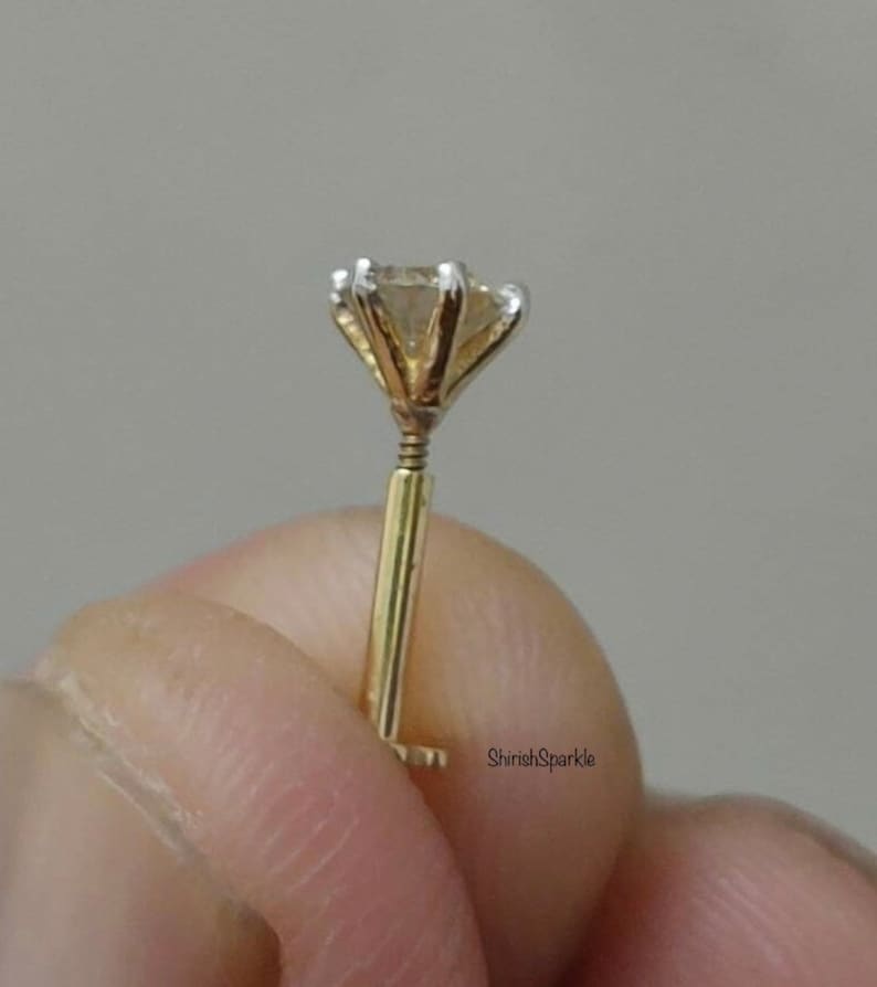 May include: A gold nose stud featuring a single round clear gemstone held in a six-prong setting. The post is straight with a threaded screw mechanism near the base of the setting. The text ShirishSparkle is visible on the lower right side.