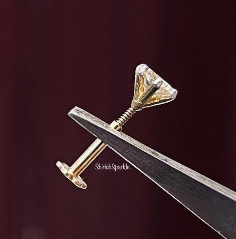 May include: A gold-toned body jewelry stud featuring a clear, faceted, triangular-cut gemstone set in a metal prong setting. The post has a threaded screw mechanism and a flat circular back disc. The text ShirishSparkle is visible on the post.