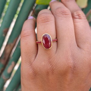 K&ouml;nnte beinhalten: Ein Goldring mit einem gro&szlig;en, ovalen, tiefroten Rubin in einer glatten Fassung. Die Ringschiene ist schmal und mit einer Reihe kleiner, runder, leuchtend pinker Steine verziert, die sich entlang der Seiten erstrecken und f&uuml;r ein detailliertes Aussehen sorgen.