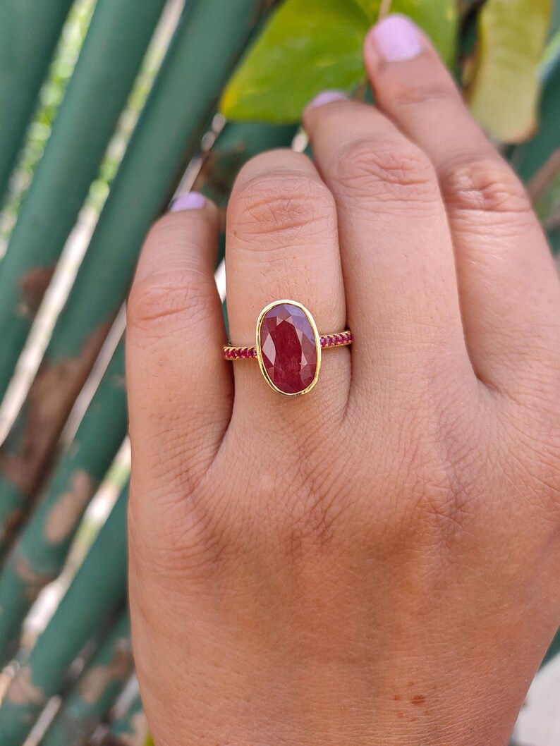 K&ouml;nnte beinhalten: Ein Goldring mit einem gro&szlig;en, ovalen, tiefroten Rubin in einer glatten Fassung. Die Ringschiene ist schmal und mit einer Reihe kleiner, runder, leuchtend pinker Steine verziert, die sich entlang der Seiten erstrecken und f&uuml;r ein detailliertes Aussehen sorgen.