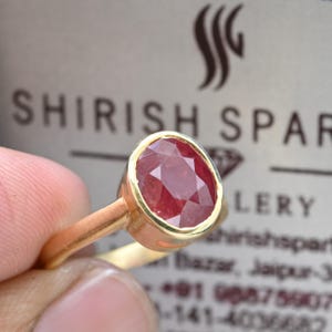 May include: A gold ring with a large, oval-cut ruby gemstone. The ruby is a deep red and set in a bezel. The ring's band is polished gold. The background shows the text "SHIRISH SPARKLERY". The ring is a piece of jewellery.