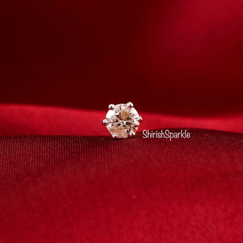 May include: A round, champagne-colored gemstone set in a six-prong silver-toned metal stud earring. The setting features a classic basket design. The text ShirishSparkle is printed in white to the right of the stud on the deep red fabric.