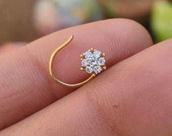 May include: A gold nose ring with a flower-shaped design. The flower is made of small, clear gemstones. The ring has a curved shape, with the gemstone flower at the end of the curve.