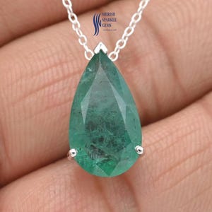 Puede incluir: Un collar de plata con un gran colgante de esmeralda facetada en forma de lágrima. La esmeralda es de color verde vibrante y está sujeta por un engaste de plata. La cadena del collar también es de plata. El colgante es el punto focal de la joya.