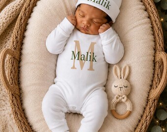 May include: A white long-sleeve onesie and matching beanie set featuring the name Malik in dark green text. The onesie includes a large tan letter M behind the name. A small, cream-colored crocheted rabbit rattle with a wooden ring is included.