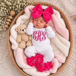 Può includere: Un body bianco a maniche lunghe per neonati con un monogramma rosa acceso sul petto, abbinato a un grande fiocco per capelli rosa acceso e scaldamuscoli con volant. Un piccolo orsacchiotto di peluche beige riposa accanto al neonato.