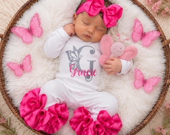 May include: A white long-sleeved infant bodysuit with a grey butterfly graphic and the name Gracie in pink script. The outfit includes a matching pink headband with a large bow and pants featuring bright pink ruffled cuffs at the ankles.