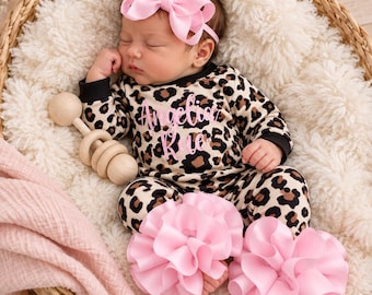 Könnte beinhalten: Ein Baby trägt ein langärmeliges Outfit mit Leopardenmuster und dem in Pink aufgedruckten Namen Angelica Kate auf der Brust. Das Baby trägt ein großes rosa Schleifen-Stirnband und rosa gerüschte Beinwärmer und hält eine Holzrassel.