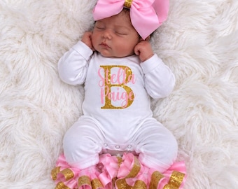 Könnte beinhalten: Ein weißer langärmeliger Baby-Body mit dem Namen Stella Paige in rosa Schreibschrift und einem großen goldenen Glitzer-Buchstaben B. Das Set enthält eine passende rosa Haarschleife und eine weiße Hose mit rosa und goldenen Glitzer-Rüschen.