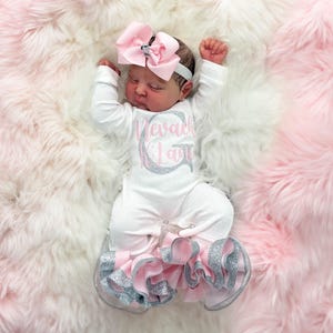 Könnte beinhalten: Ein Neugeborenes in einem weißen Strampler mit dem Namen "Nevaeh Kila" in rosa und silbernem Glitzer. Der Strampler hat rosa und silberne Rüschen. Das Baby trägt ein rosa Schleifen-Stirnband und liegt auf einer rosa-weißen, flauschigen Decke.