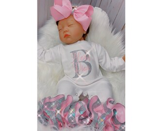 May include: A white baby onesie with a silver glitter letter B and the text "Bloom" in pink glitter. The onesie is paired with a pink and silver glitter ruffle skirt. The baby is wearing a pink bow headband.