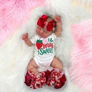 Op de afbeelding: Een pasgeboren baby draagt een witte romper met een aardbei-afbeelding en de tekst "So Berry Sweet". De baby draagt ook een grote rode strik en gefranjerde beenwarmers met een aardbeienpatroon. De baby ligt op een zachte witte en roze vloerkleed.