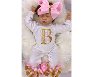 May include: A white baby onesie with pink and gold glitter lettering that says "Stella Paige" and a gold glitter letter "B". The baby is wearing a pink bow headband and pink and gold ruffled shoes.