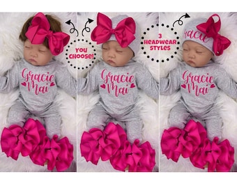 Könnte beinhalten: Ein grauer Baby-Strampler mit dem Text "Gracie Mai" in rosa Schrift und drei rosa Herz-Akzenten. Der Strampler wird mit einem rosa Rüschenrock und einer Auswahl von drei rosa Haarbändern kombiniert: einer Schleife, einem Haarreif mit Schleife oder einer Mütze mit Schleife.