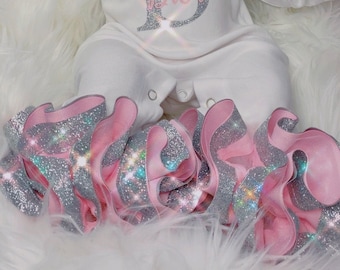 May include: A white baby onesie with the letter "D" in silver glitter and the word "Dior" in pink glitter. The onesie is paired with a pink and silver glitter ruffle skirt.
