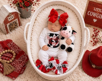 May include: A baby wearing a white long-sleeve onesie with a black and white cow print letter B and the name Brooklyn Paige in red script. The baby wears a large red hair bow and ruffled leg warmers, holding a small plush cow toy in a white basket.