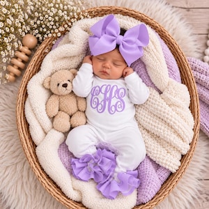 May include: A sleeping infant wearing a white long-sleeve onesie with a purple monogrammed design on the chest, a large purple hair bow, and purple ruffled leg warmers. A small tan plush teddy bear rests beside the infant in a woven basket.