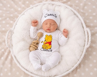 May include: A white long-sleeved baby onesie and matching hat featuring a graphic of Winnie the Pooh and the name Charlie printed in black. The infant is holding a wooden rattle while resting inside a white woven basket with a soft lining.