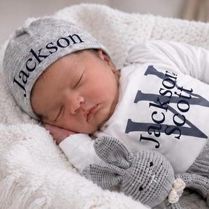 May include: A gray beanie with the name Jackson printed in black, paired with a white long-sleeve shirt featuring the name Jackson Scott in large black letters, and a small gray crocheted rabbit toy with a white bow detail.