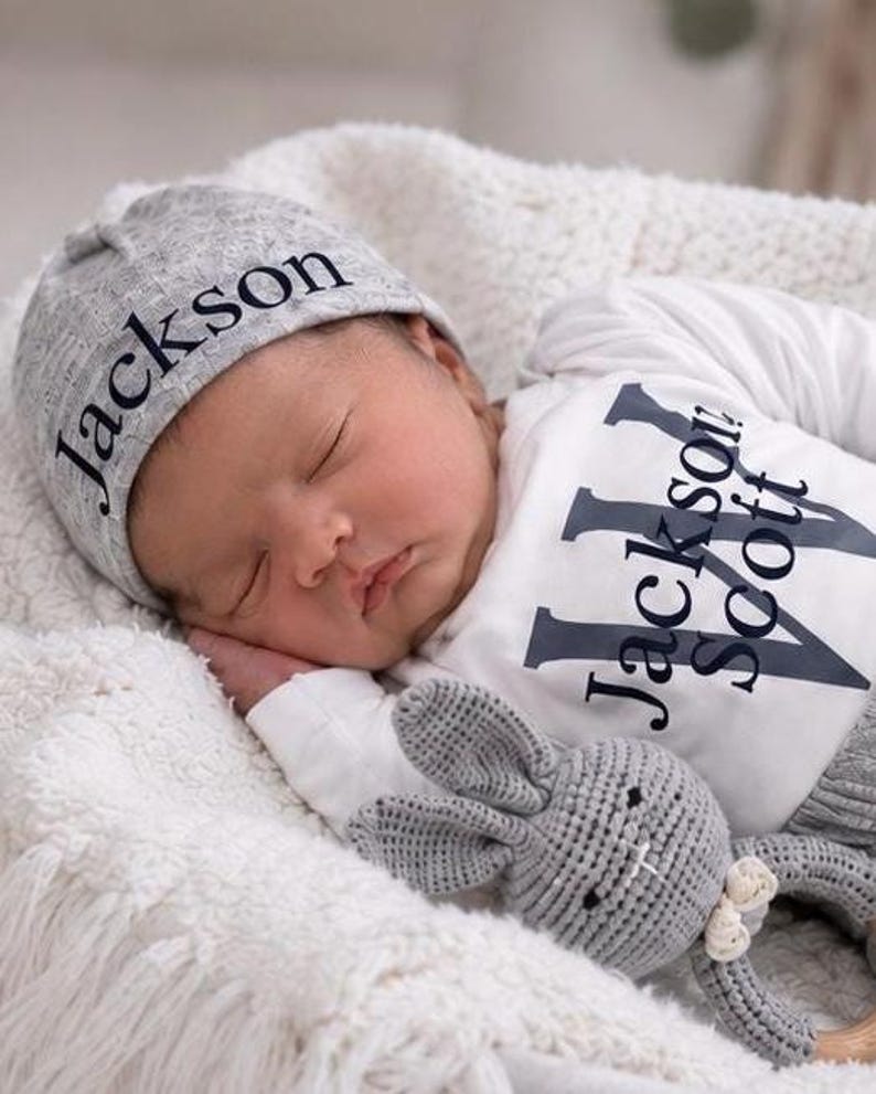 May include: A gray beanie with the name Jackson printed in black, paired with a white long-sleeve shirt featuring the name Jackson Scott in large black letters, and a small gray crocheted rabbit toy with a white bow detail.