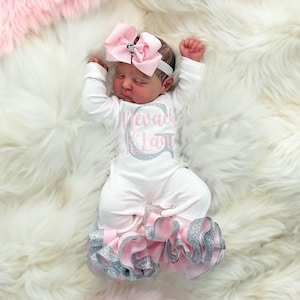 May include: A baby wearing a white onesie with the name "Nevaeh Kila" in silver glitter lettering. The onesie has pink and silver ruffled trim at the feet. The baby is lying on a white fluffy blanket with a pink fluffy blanket in the background. A pink bow headband is on the baby's head.