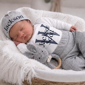 May include: A newborn baby wearing a grey knit hat with the name Jackson embroidered in black, a white long-sleeve bodysuit with the name Jackson Scott printed in black, and grey knit pants, holding a grey crocheted rabbit rattle with a wooden ring.