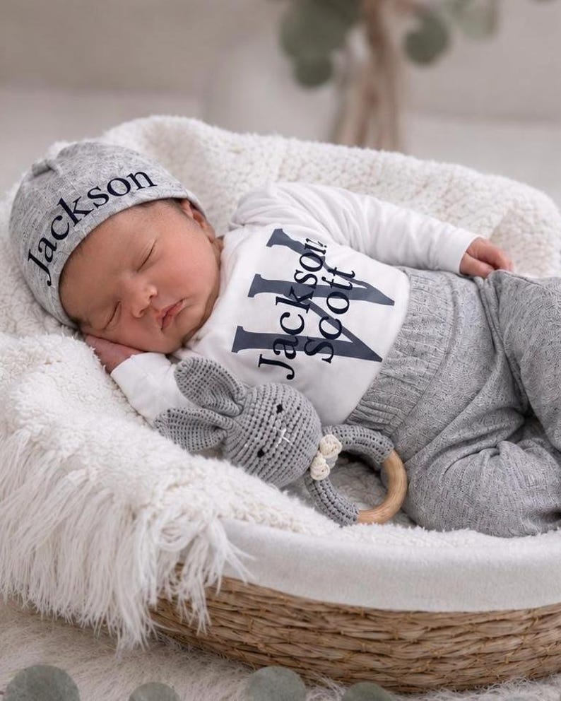 May include: A newborn baby wearing a grey knit hat with the name Jackson embroidered in black, a white long-sleeve bodysuit with the name Jackson Scott printed in black, and grey knit pants, holding a grey crocheted rabbit rattle with a wooden ring.
