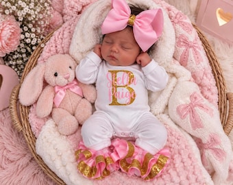 May include: A sleeping infant wearing a white long-sleeve bodysuit with the name Stella Paige printed in gold glitter and a large pink hair bow. The infant is dressed in white ruffled trousers with gold glitter trim and sits beside a plush pink bunny.