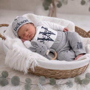 May include: A newborn baby wearing a grey beanie with the name Jackson, a white long-sleeve bodysuit with the name Jackson Scott, and grey pants with the initials JW embroidered on the leg, holding a grey crocheted rabbit toy with a wooden ring.