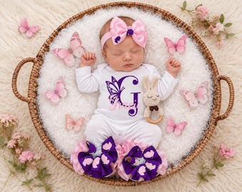 May include: A white long-sleeve bodysuit featuring a purple butterfly wing design integrated into the letter G and the name Gracie in pink script. The outfit includes a pink bow headband, purple and pink ruffled leg warmers, and a crochet bunny rattle.