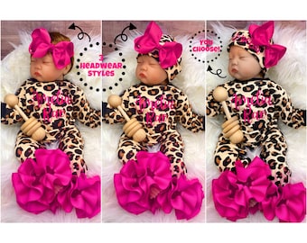 May include: Three baby outfits with a leopard print design, each featuring a different headwear style. The outfits include a long-sleeved onesie with a ruffled bottom and a matching bow or hat in a vibrant pink. Each outfit includes a wooden rattle.
