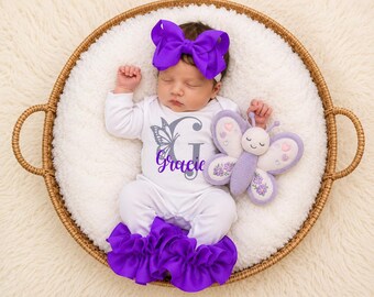 May include: A white long-sleeve onesie with a grey butterfly graphic and the name Gracie in purple script, paired with a large purple hair bow and purple ruffled leg warmers, accompanied by a plush purple and white butterfly toy.