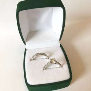 May include: A pair of diamond rings in a green velvet ring box. The engagement ring has a large center stone and a band of smaller diamonds. The wedding band is a simple band with small diamonds.